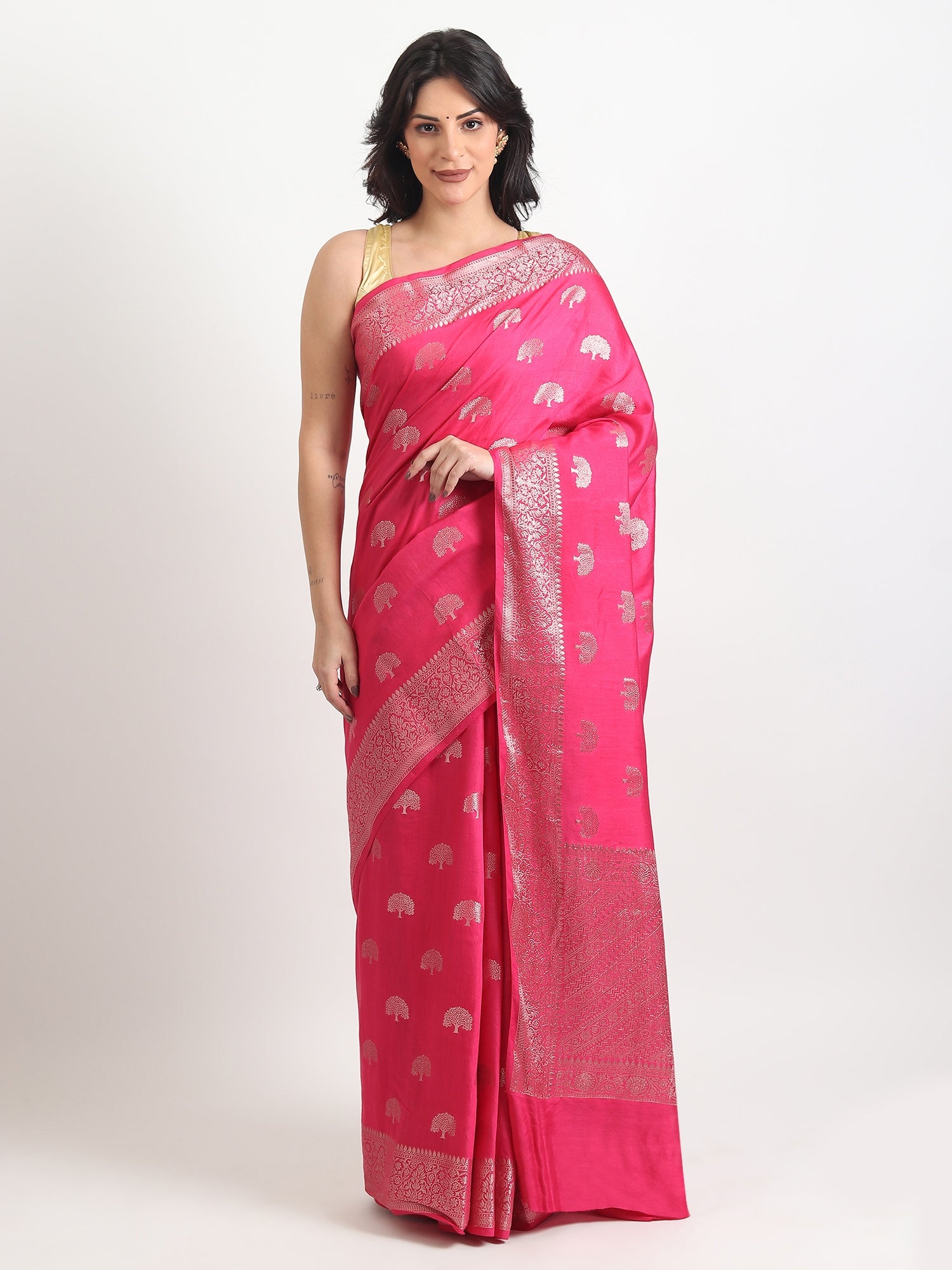 A woman with dark hair stands against a plain white background, wearing a vibrant rani pink Banarasi silk-blend saree. The saree features a smooth, glossy drape and is adorned throughout with small, evenly spaced woven silver tree motifs (butas). It includes a thick, intricately woven silver zari border and pallu. She has styled it with a sleeveless metallic gold blouse and has subtle makeup on, presenting a classic ethnic look.