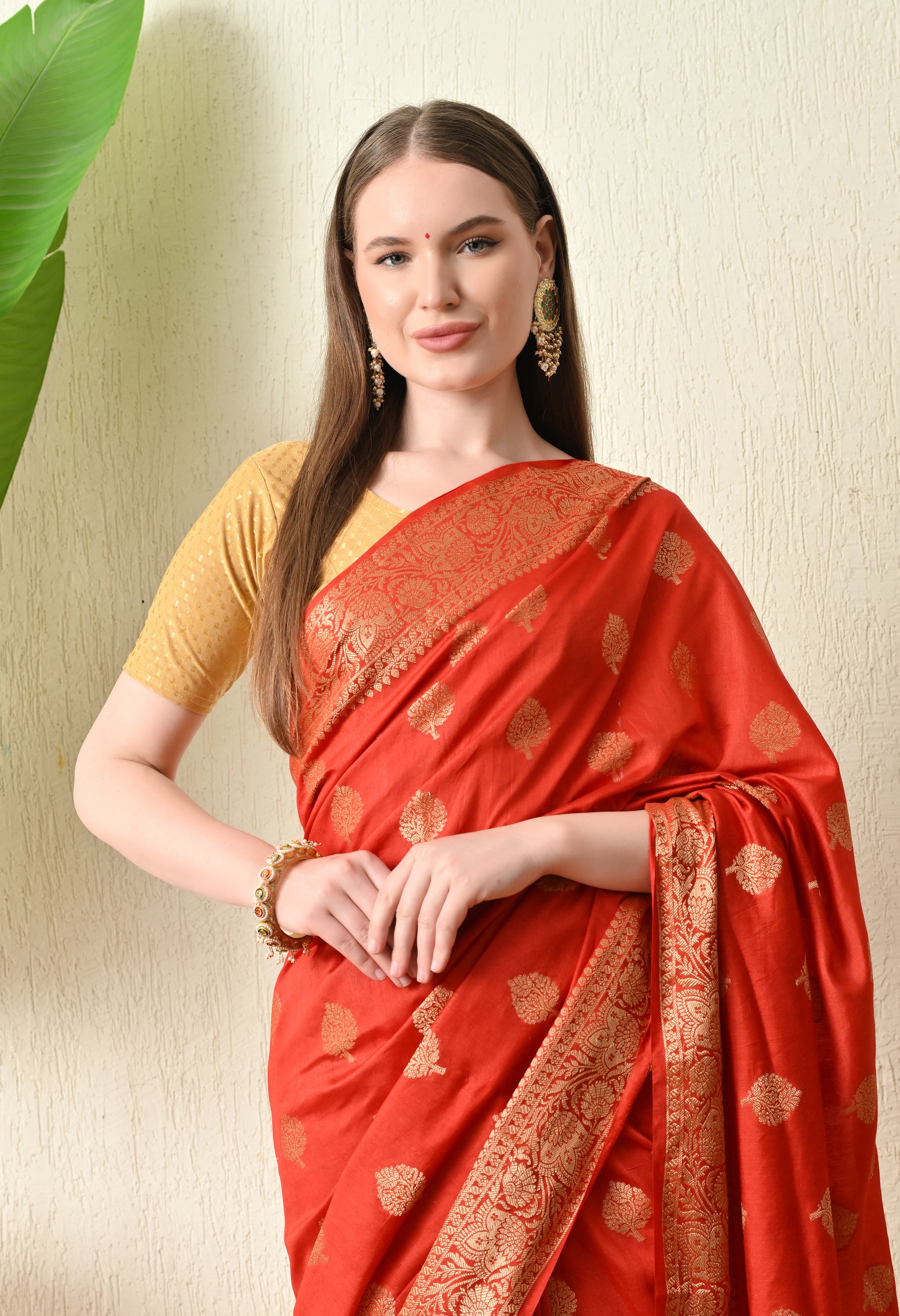A close-up shot of the same red Banarasi wedding saree, highlighting the fine detail of the gold zari border and the delicate leaf patterns. The woman is accessorized with traditional gold jhumka earrings, a bangle, and a small red bindi. The gold silk blouse provides a rich contrast to the red fabric, emphasizing the saree's luxurious texture.