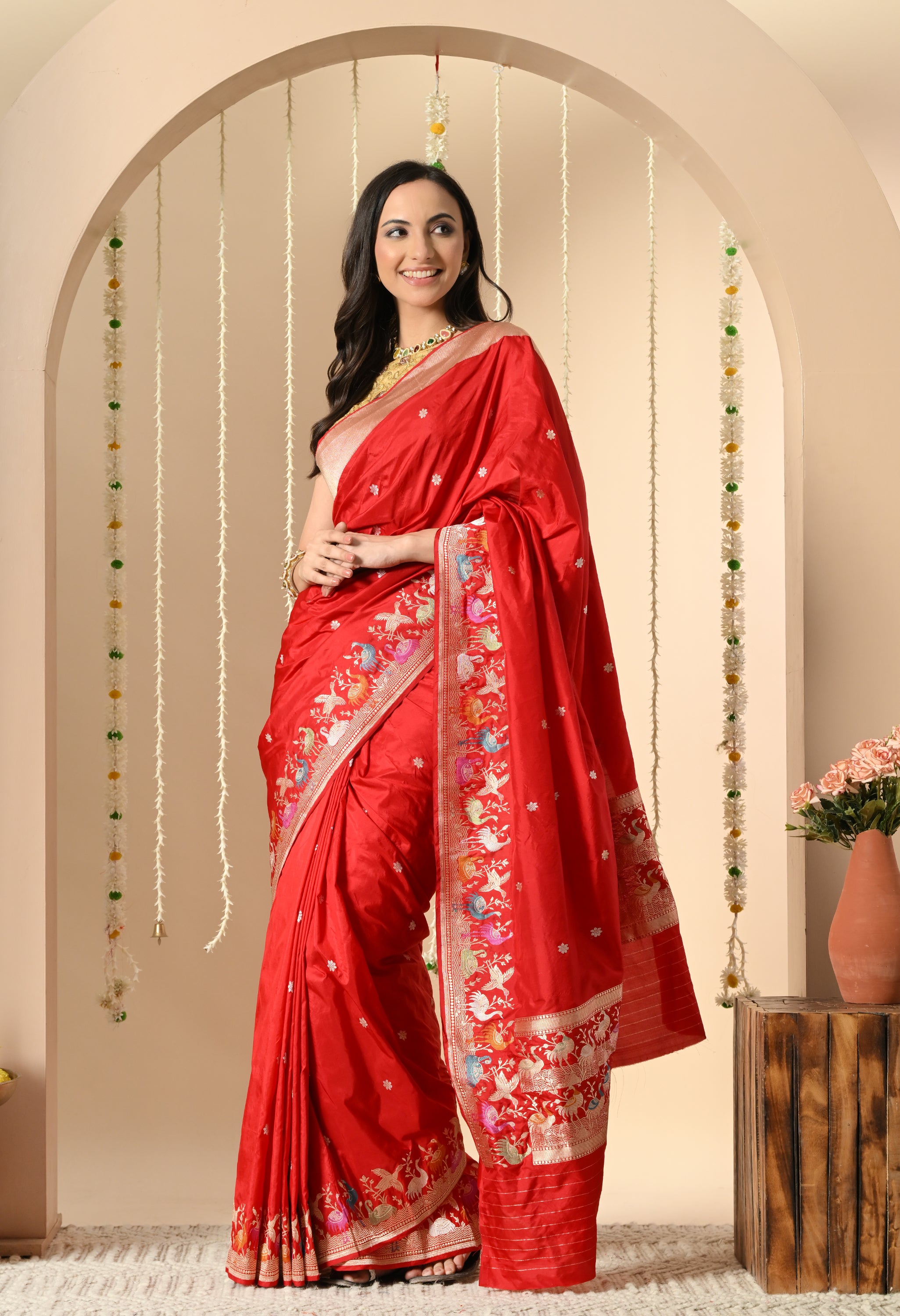 A beautiful woman models a luxurious vibrant red Katan silk saree from The Weavers Shop by Raavi. The Silk Mark certified saree body is embellished with subtle woven Zari motifs (butis). The wide, multicolored Shikarga border showcases intricate Zari work depicting scenes of animals and human figures, which also extends to the rich pallu. She is accessorized with a gold blouse and traditional jewelry, and is photographed from the side in a cream-colored arched setting.