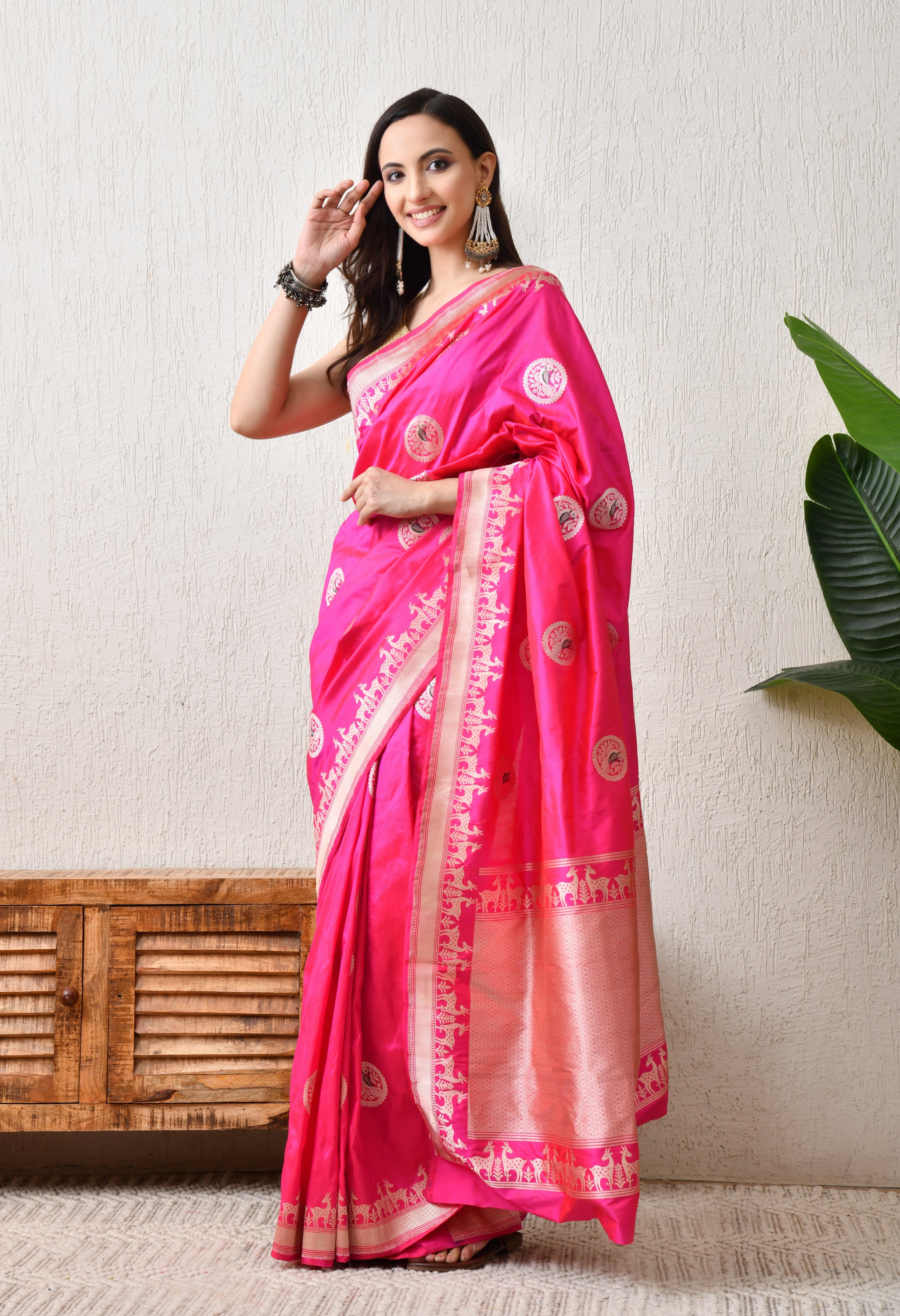 A side-angle view of a woman posing in a vibrant Barbie pink Hand Woven Katan Silk saree from The Weavers Shop by Raavi. This profile shot highlights the elegant drape and the rich texture of the Silk Mark Certified fabric. The intricate silver zari Shikarga border with its signature deer motifs is clearly visible along the length of the saree, complemented by circular bird-design butis. She is styled with a sleeveless gold blouse and traditional dangling earrings, set against a neutral, minimalist interior