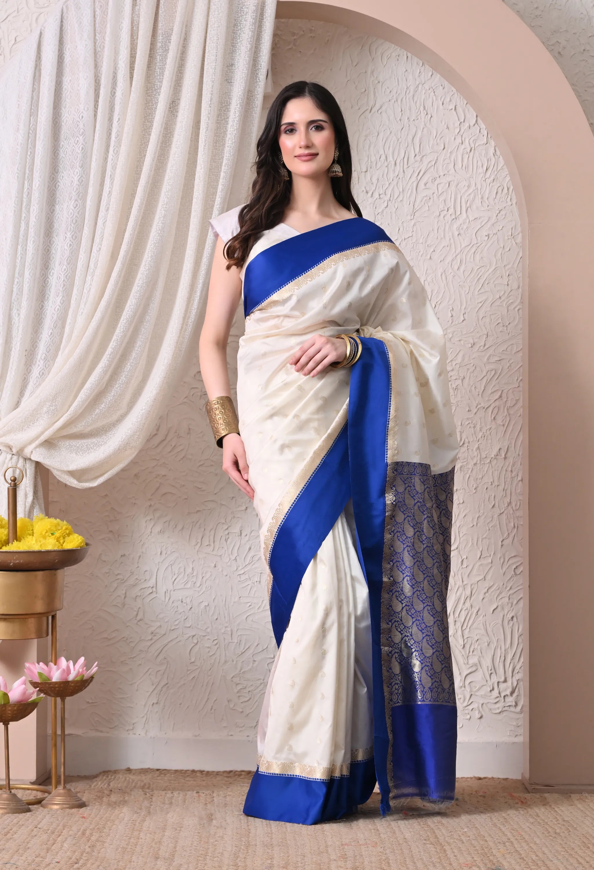 A woman posing in an elegant off-white silk blend saree featuring a striking royal blue border and a richly detailed paisley-patterned pallu.
