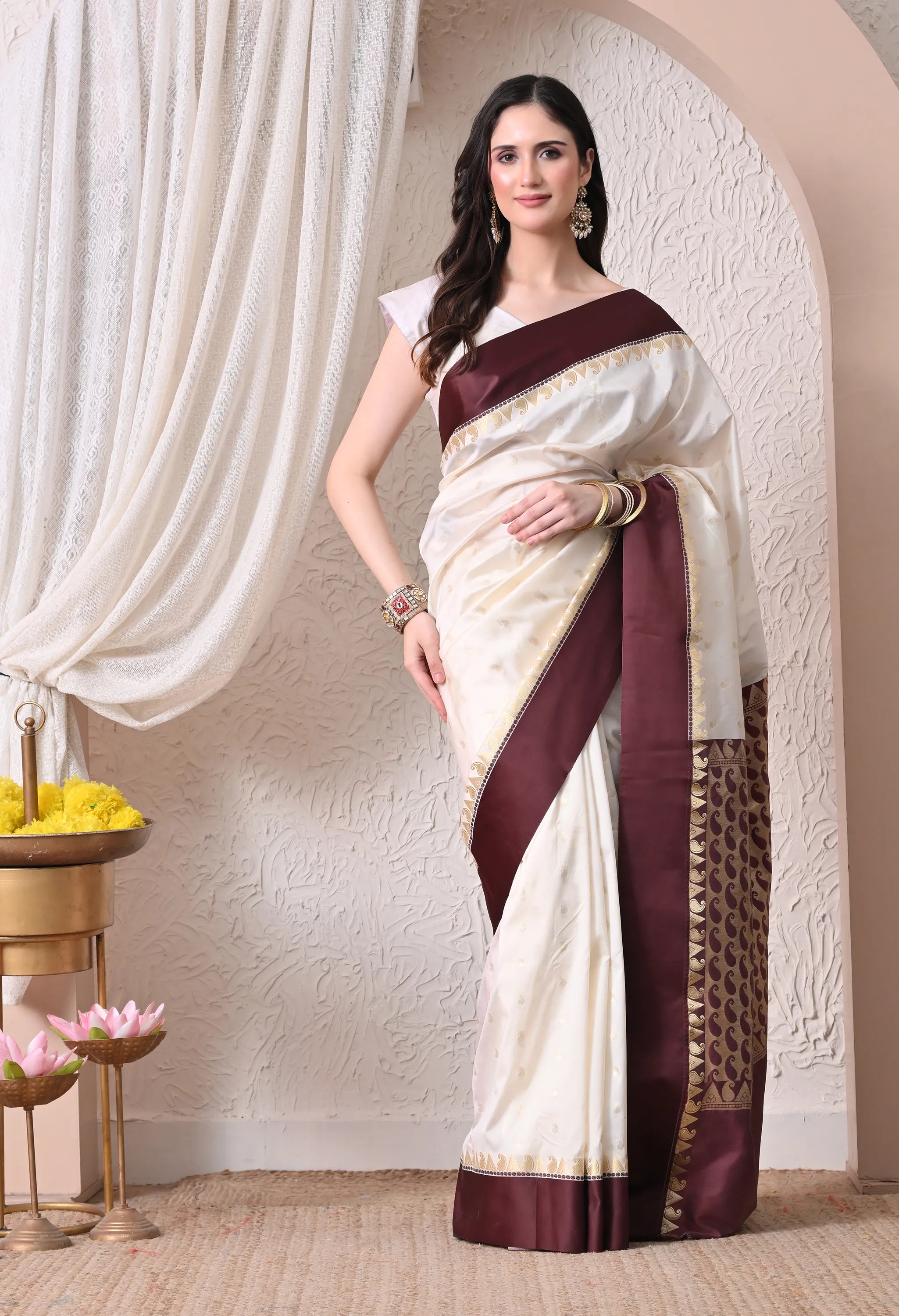A woman posing in an elegant off-white cotton saree featuring a deep maroon border with golden zari details and a matching paisley-patterned pallu.
