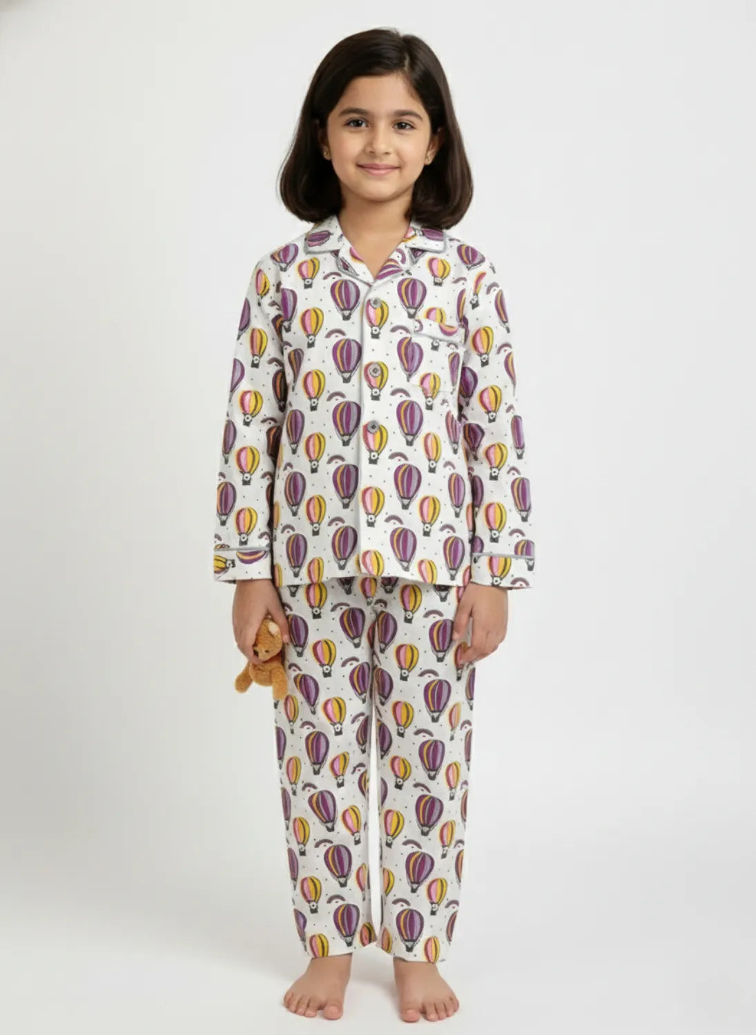 A young girl standing against a white background, wearing a white long-sleeved button-up pajama set with a colorful hot air balloon print.