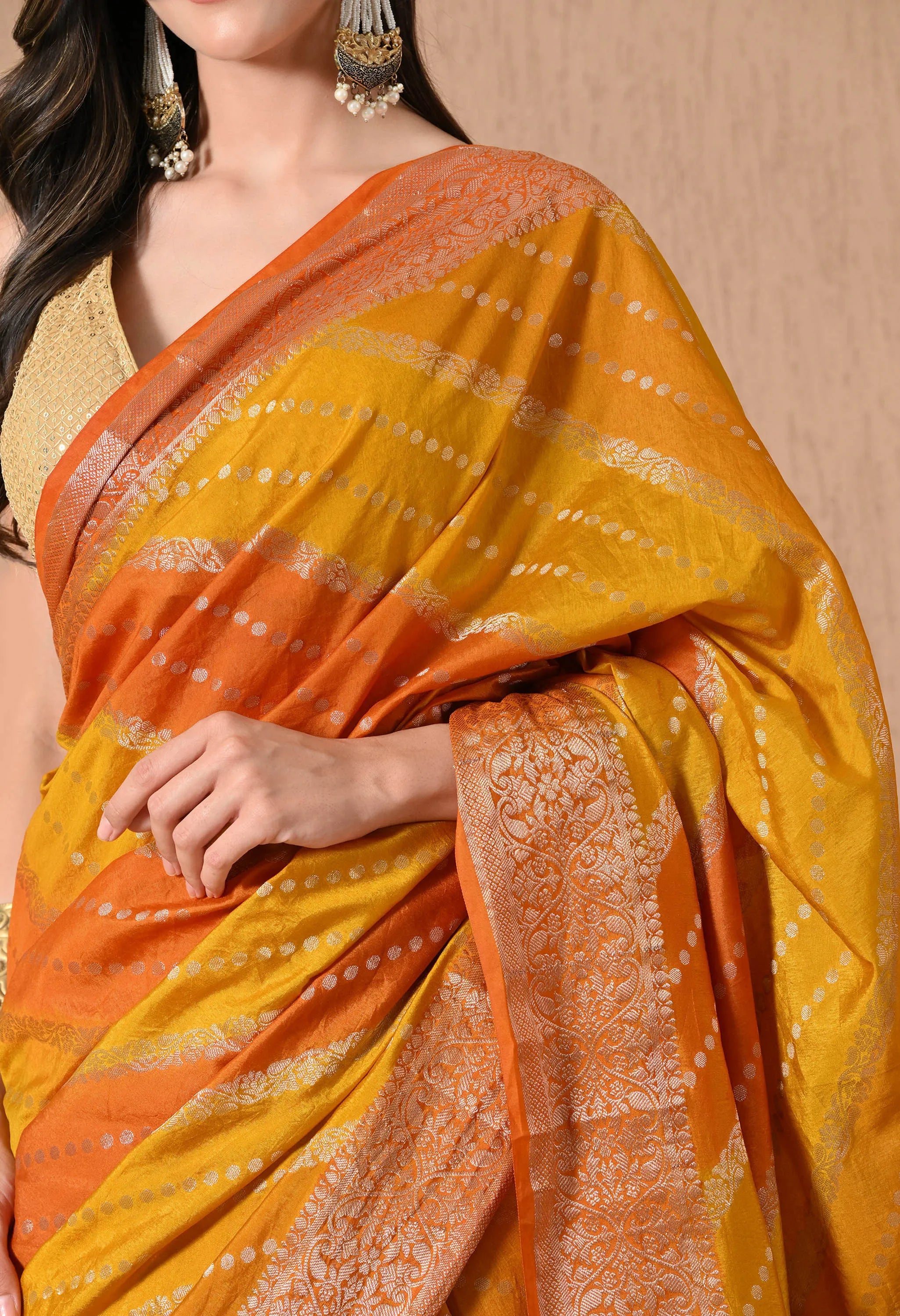 A close-up shot of a model showcasing a Rangkat Banarasi silk saree from The Weavers Shop, focusing on the intricate textures of the fabric. The image highlights the golden zari work on the borders and the transition of the Rangkat diagonal stripes in vibrant mustard yellow and deep orange. Small, silver-toned woven dots (buttis) are scattered across the body, while the model is styled with a sleeveless gold  blouse and traditional statement jewelry.