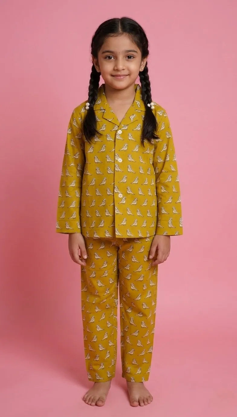 young girl with braided hair wearing a mustard yellow 100% cotton pajama set against a soft pink background. The night suit features a long-sleeved button-down shirt with a white notched collar and matching straight-leg trousers, both decorated with a delicate white bird or animal print.