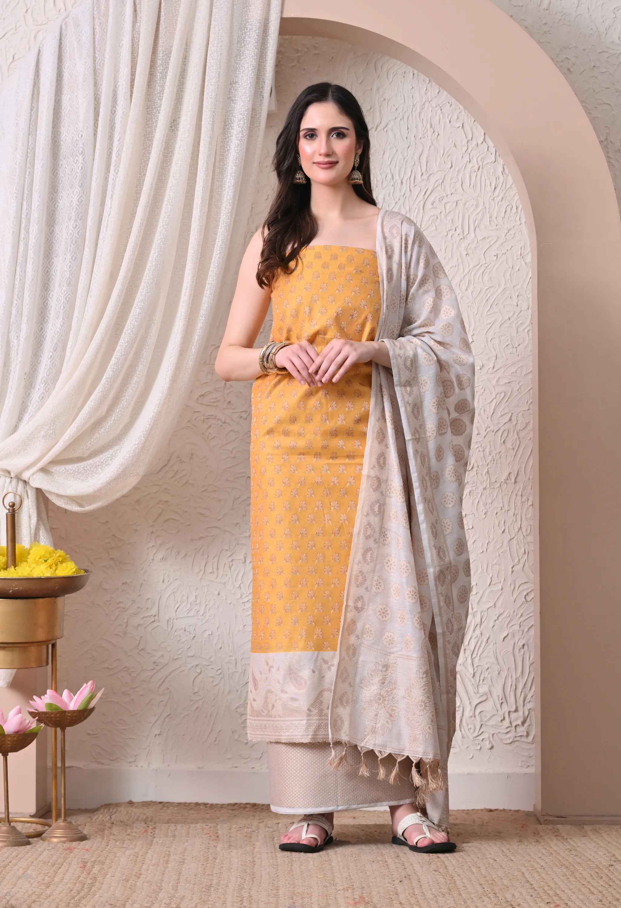 A woman posing in a vibrant mustard yellow and off-white woven silk salwar suit set featuring detailed ethnic motifs, an off-white paisley border, and a matching tasseled dupatta.
