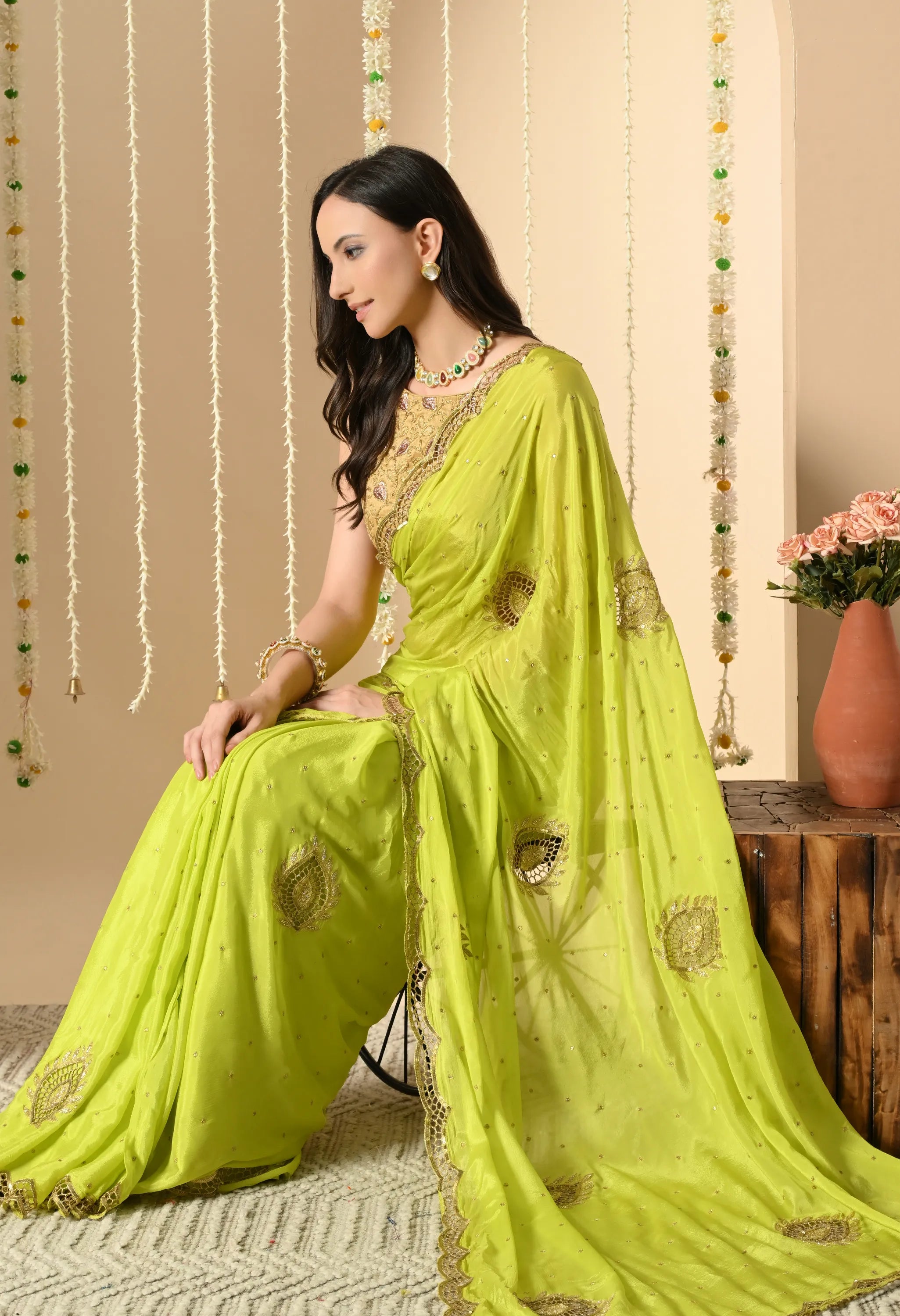 A model seated in a vibrant lime-green chiffon saree by The Weavers Shop by Raavi, displaying the detailed gold zardozi motifs and cut-work in drop motif and scalloped edge. She is paired with a gold blouse and traditional jewellery, set against a festive backdrop with floral garlands.