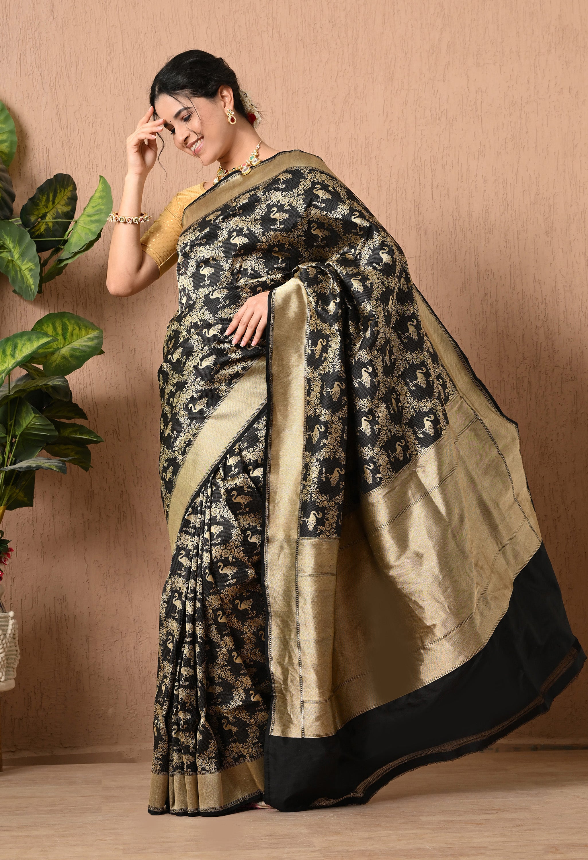 A woman stands in a profile view, showcasing a sophisticated black handwoven  pure Katan Silk saree from The Weavers Shop by Raavi. The fabric is adorned with an intricate hand-woven zari Shikarga design, featuring detailed gold patterns of animals and nature. This Silk Mark Certified saree includes a wide, shimmering gold border that flows elegantly as she poses. She is wearing a gold blouse and ornate traditional jewelry, with her hair styled in a bun.
